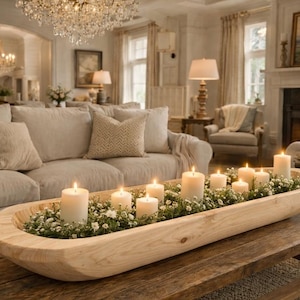 May include: A living room scene with a light-colored sofa, pillows, and a wooden coffee table. A wooden trough filled with greenery and lit candles sits on the table. A chandelier hangs above.