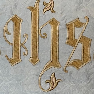 May include: Close-up of an embroidered design featuring the letters IHS in gold thread. The letters are in a decorative, ornate font, set against a light blue fabric with a subtle floral pattern. The embroidery is detailed and elegant.