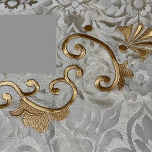 May include: Close-up of a cream-colored fabric with an embossed floral pattern. Gold-colored, ornate embroidery in a scrolling design adorns the fabric. The embroidery features a stylized floral motif and adds a touch of elegance.