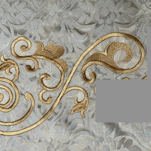 May include: Close-up of a gold embroidered floral design on a light gray fabric. The embroidery features swirling lines, leaves, and a textured pattern. The background fabric has a subtle floral pattern.