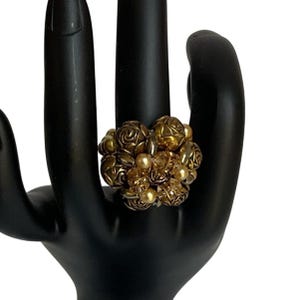 May include: A gold-toned statement ring featuring a cluster of detailed floral and geometric shapes, accented with small, round, pearl-like beads. The ring is displayed on a black hand-shaped jewelry display, showcasing its intricate design and vintage aesthetic.