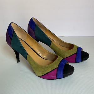 May include: A pair of open-toe high heels with a color block design. The heels feature a combination of green, purple, blue, and black suede panels. The heels have a stiletto heel.