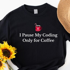 May include: Black t-shirt with the text "I Pause My Coding Only for Coffee" in white. The shirt features a red coffee mug graphic with a candy cane and snowflake design. A straw hat and sunflowers are in the background.