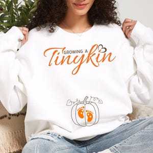 May include: White sweatshirt with the text "Growing a Tinykin" in orange script. Below the text is a pumpkin graphic with orange baby footprints. The sweatshirt is a maternity top.