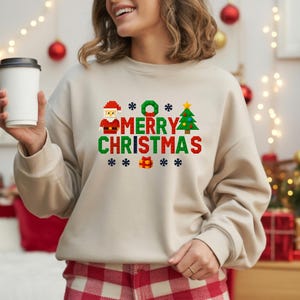 May include: Beige sweatshirt with a pixelated "MERRY CHRISTMAS" design in red, green, and white. The design includes Santa, a Christmas tree, and gift boxes. The person is holding a white coffee cup.