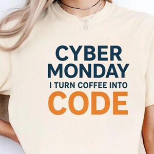 May include: Cream-colored t-shirt with the text "CYBER MONDAY" in navy blue and "CODE" in orange. The phrase "I TURN COFFEE INTO" is in between the two phrases. The shirt is a casual wear item.