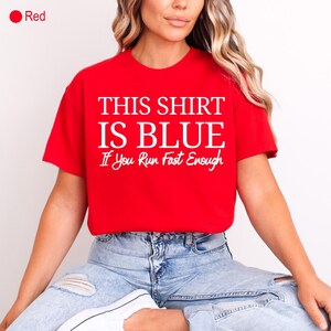 May include: A red t-shirt with the text "THIS SHIRT IS BLUE If You Run Fast Enough" in white. The shirt is short-sleeved and is paired with light blue jeans.