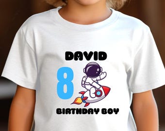 May include: White t-shirt with a colorful graphic for a birthday. The design includes the name "DAVID" in black, the number "8" in blue, and an astronaut on a rocket. The text "BIRTHDAY BOY" is also included.