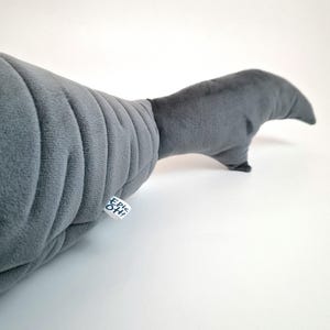 May include: A gray plush toy shaped like a whale's tail. The plush has a ribbed texture on the body and a smooth tail fin. A small tag with the text "Epic Oh!" is attached to the body.