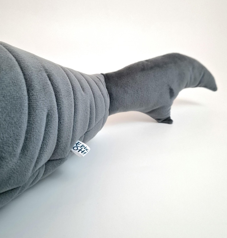 May include: A gray plush toy shaped like a whale's tail. The plush has a ribbed texture on the body and a smooth tail fin. A small tag with the text "Epic Oh!" is attached to the body.