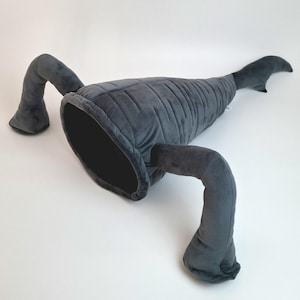 May include: A gray plush cat bed shaped like a whale, with a large opening and two curved arm-like appendages. The whale-shaped bed is made of soft, velvety material and is designed for pets to snuggle in.