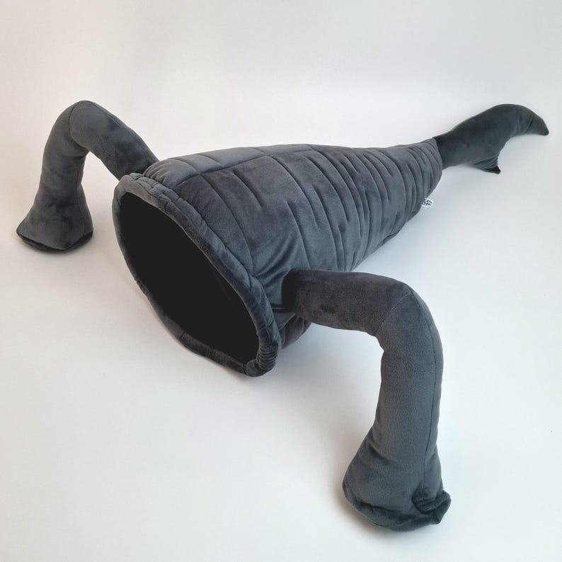 May include: A gray plush cat bed shaped like a whale, with a large opening and two curved arm-like appendages. The whale-shaped bed is made of soft, velvety material and is designed for pets to snuggle in.