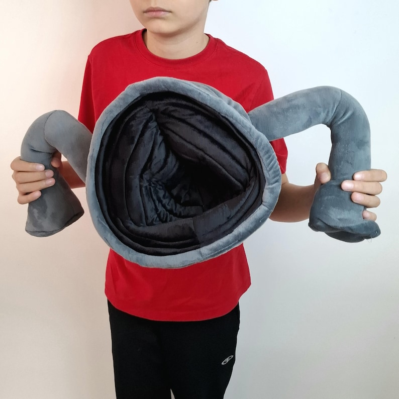 May include: A gray plush toy shaped like a large, open-mouthed creature with two curved arms. The interior is black, and the exterior is a soft gray fabric. The toy is held up by a person wearing a red shirt and black pants.
