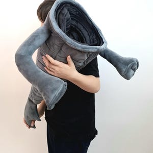May include: A large, gray plush toy shaped like a stylized creature, held by a person. The toy has a quilted body, a wide opening, and two long, arm-like appendages. The plush is a soft, velvety material.