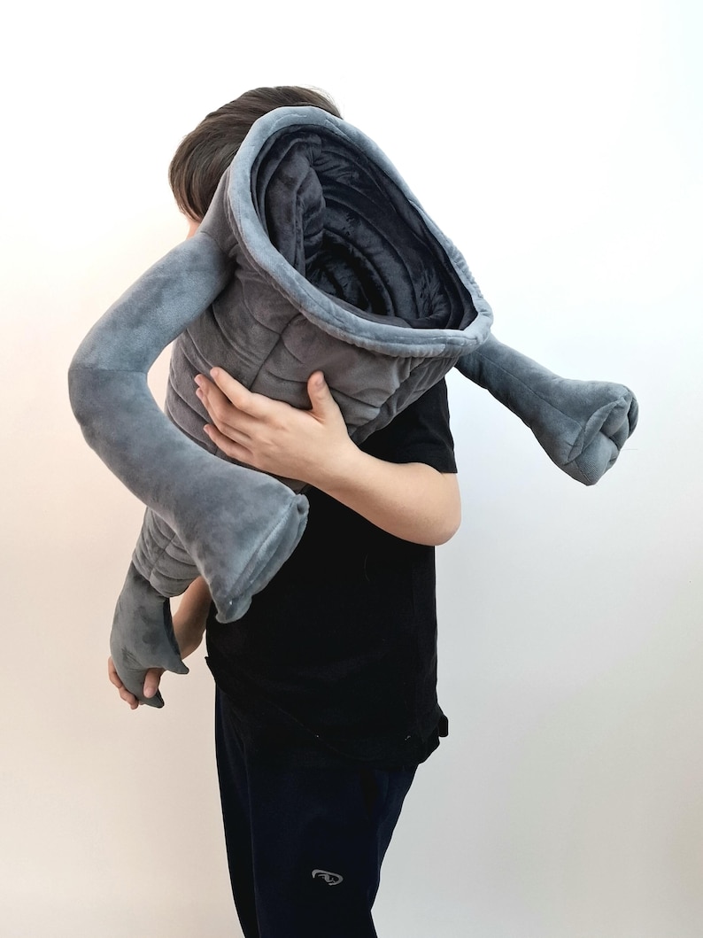 May include: A large, gray plush toy shaped like a stylized creature, held by a person. The toy has a quilted body, a wide opening, and two long, arm-like appendages. The plush is a soft, velvety material.