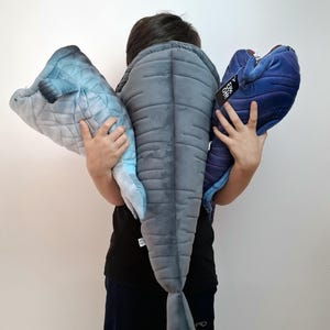 May include: A person holding three plush toy fish. The toys are in shades of blue and gray, with quilted and textured details. One toy has a tag that reads "A Epic Oh!". The toys are soft and cuddly.