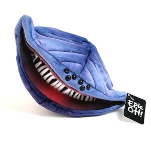 May include: A plush, blue, leaf-shaped creature with a wide, toothy grin. The plush has a quilted texture and several black button-like eyes. The inside of the mouth is red, and the teeth are white. A black tag with the text "Epic OH!" is attached.