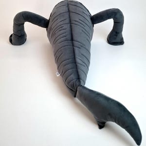 May include: A gray plush toy shaped like a sea creature, featuring a segmented body, two arms, and a tail. The toy is made of soft fabric and has a tag attached. The plush is designed to resemble a stylized aquatic animal.