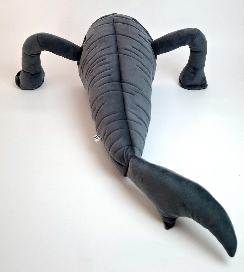 May include: A gray plush toy shaped like a sea creature, featuring a segmented body, two arms, and a tail. The toy is made of soft fabric and has a tag attached. The plush is designed to resemble a stylized aquatic animal.