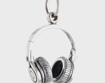 May include: A silver-colored pendant in the shape of headphones hangs from a black cord. The headphones have detailed earcups and a curved headband. The pendant is designed to resemble over-ear headphones, perfect for music lovers.
