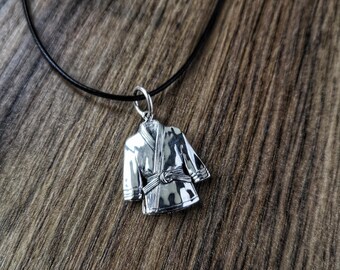 May include: A silver-colored pendant necklace featuring a miniature karate gi, or uniform, with a tied belt. The pendant hangs from a thin, black cord. The background is a textured, brown surface.