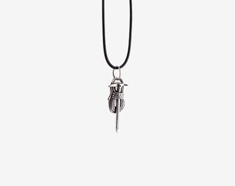 May include: A silver-colored pendant necklace featuring a stylized, detailed design. The pendant is suspended from a black cord. The pendant has a central, vertical element with a wrapped design and a pointed tip.