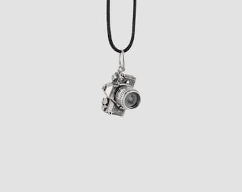 May include: A silver-colored camera pendant hanging from a black cord. The pendant is a detailed miniature replica of a camera, with a lens, buttons, and other features. The background is a solid, light gray color.