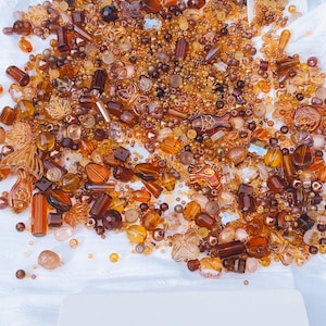 May include: A selection of amber and brown beads in diverse shapes, including round, rectangular, and floral designs. The beads are spread on a white surface, with a card at the bottom reading "L'Atelier Jardin de Coco."