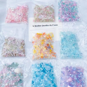 Puede incluir: Nueve pequeñas bolsas de plástico transparente llenas de cuentas y adornos coloridos. Las bolsas contienen varios colores, incluyendo rosa, verde, azul y morado. Una etiqueta blanca con el texto "L'Atelier Jardin de Coco" es visible.