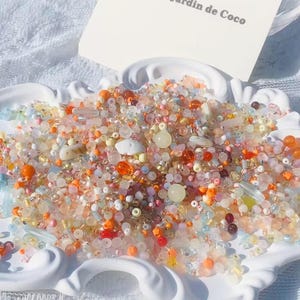 May include: A white, decorative ceramic dish filled with a vibrant mix of beads in shades of orange, pink, yellow, and clear. Some larger, round beads are also present. The dish holds a card with the text "L'Atelier Jardin de Coco".