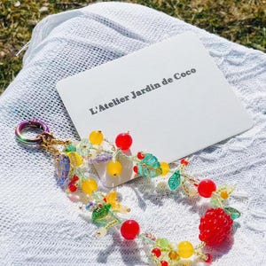 May include: A beaded keychain with a fruit and leaf design. The keychain features red, yellow, and green beads, with a large red raspberry and a card that reads "L'Atelier Jardin de Coco".