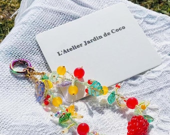 May include: A colorful beaded keychain with a fruit and leaf design. The keychain features red, yellow, and green beads, with a large red raspberry and a card that reads "L'Atelier Jardin de Coco".