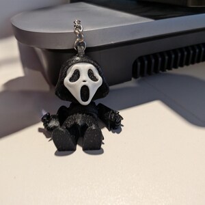 Peut inclure: Un porte-clés représentant le personnage du film Scream, en noir et blanc. Le porte-clés représente une petite figurine du tueur Ghostface avec un masque blanc et une robe noire. La figurine tient un petit couteau. Le porte-clés est attaché à une chaîne argentée.