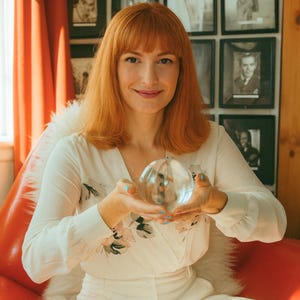 May include: A clear glass sphere held in both hands. The sphere reflects the surrounding environment. The person is wearing a white dress with floral accents. The background includes framed black and white photographs and a red chair.