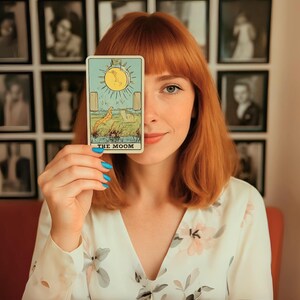May include: A tarot card with a yellow sun and the text "THE MOOM" is held in front of a person's face. The card features a landscape with a yellow sun, a dog, and two pillars. The person has red hair and is wearing a floral top.