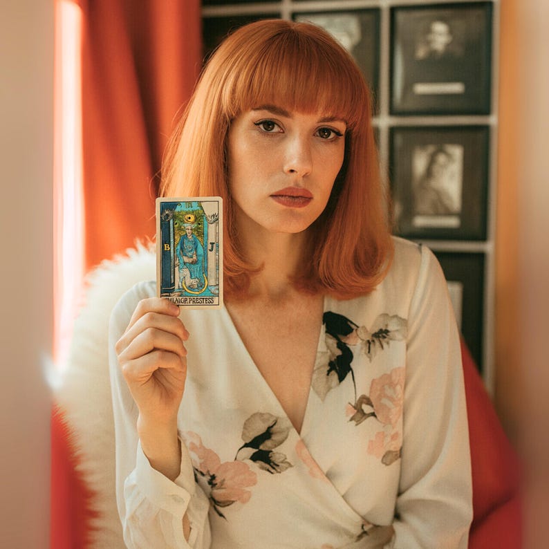 May include: A woman holding a tarot card labeled "The High Priestess." The card features a blue-robed figure with a golden crown. The woman wears a white floral print dress and has red hair.