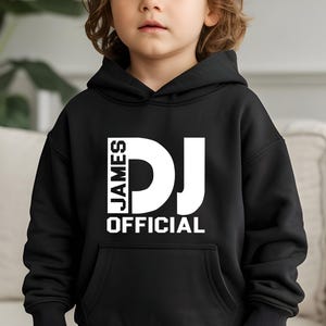 Könnte beinhalten: Ein schwarzer Kapuzenpullover mit einem weißen Grafikdruck auf der Brust. Der Text JAMES steht vertikal neben einem großen DJ, wobei das Wort OFFICIAL horizontal darunter gedruckt ist. Das Kleidungsstück verfügt über eine Kängurutasche und gerippte Bündchen.