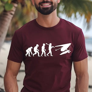 May include: A maroon short-sleeved t-shirt featuring a white graphic print that depicts the stages of human evolution, starting with a primate and ending with a person flying a hang glider, all arranged in a horizontal line across the chest.