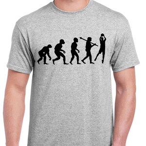 May include: A light grey t-shirt with a black graphic illustrating human evolution, ending with a basketball player. The design shows a sequence of figures evolving into a person shooting a basketball. The shirt is made of a soft material.