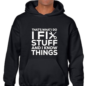 Op de afbeelding: Zwarte hoodie met de witte tekst "THAT'S WHAT I DO I FIX STUFF AND I KNOW THINGS". Het ontwerp bevat een moersleutel en een schaar. De hoodie heeft een voorzak en een capuchon met trekkoord.