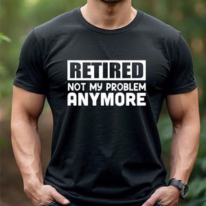 Puede incluir: Una camiseta negra de manga corta con texto blanco impreso en el pecho que dice "RETIRED NOT MY PROBLEM ANYMORE" en una fuente negrita sin remates. La camiseta tiene un cuello redondo clásico y un corte estándar de tela negra.