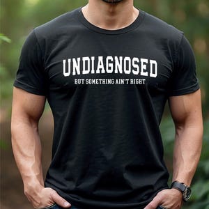 Op de afbeelding: Zwart T-shirt met de witte tekst "UNDIAGNOSED" boven "BUT SOMETHING AIN'T RIGHT". Het shirt heeft korte mouwen en lijkt van een zacht materiaal te zijn. De persoon draagt een zwart horloge.