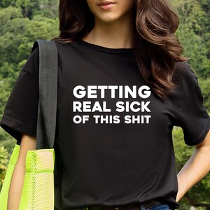 Op de afbeelding: Zwart T-shirt met de witte tekst "GETTING REAL SICK OF THIS SHIT". Het shirt heeft een ronde hals en wordt gedragen met een blauwe spijkerbroek. Een zwarte band en een neon groene tas zijn zichtbaar.