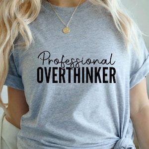 May include: A heather grey short-sleeve t-shirt featuring the text Professional OVERTHINKER printed in black. The word Professional is in a cursive script font, while OVERTHINKER is in a bold, sans-serif capital letter font.