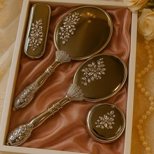 Könnte beinhalten: Ein Vintage-Kosmetikset in einer weißen Box mit einem Handspiegel, einem Kompaktspiegel, einer Bürste und einem Lippenstiftetui. Das Set hat silberfarbene Griffe und Rahmen mit floralen und Kristallverzierungen. Das Set wird auf einem rosa Satinstoff präsentiert.