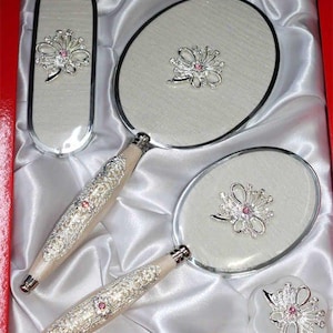 以下が含まれることがあります： 赤い箱に入った手鏡、コンパクトミラー、ブラシ、小さな容器のバニティセット。セットは、明るい色の背景にピンクのアクセントが付いた銀色の花の装飾が特徴です。