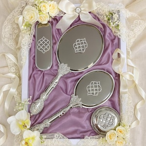 May include: A silver vanity set comprising a hand mirror, a small oval mirror, a compact, and a round container. Each item has a diamond-like pattern. The set is presented on purple fabric, with white flowers and ribbons as decoration.