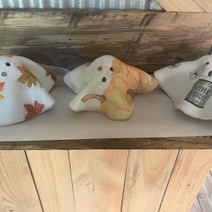 May include: Three white ceramic ghost figurines with different Halloween-themed designs. One ghost has autumn leaves, another has a pumpkin, and the third has a skull and the text "Spider Venom".