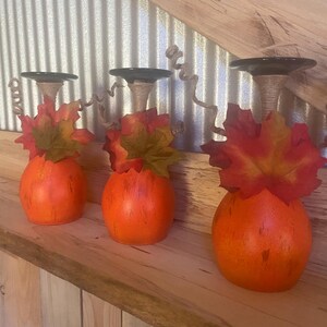 May include: Three orange pumpkin-shaped candle holders with black tops and fall leaf accents. The candle holders are displayed on a wooden shelf against a corrugated metal and wood background.