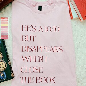 May include: A light pink t-shirt featuring dark red text that reads: HE'S A 10/10 BUT DISAPPEARS WHEN I CLOSE THE BOOK. The shirt has a crew neckline and short sleeves, with the printed message centered on the front of the garment in a serif font.